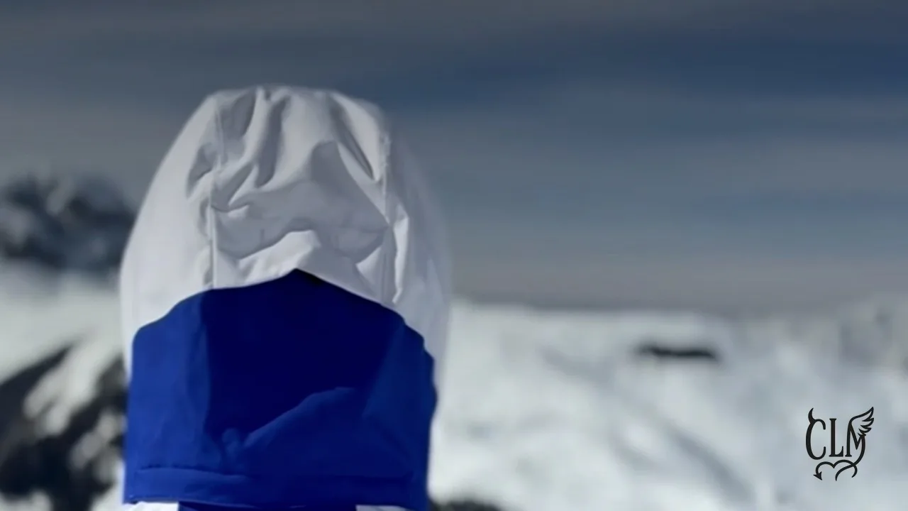 Image du clip Comment quitter le froid ? : une silhouette encapuchonnée de blanc et de bleu, de dos, devant une étendue de montagnes enneigées.