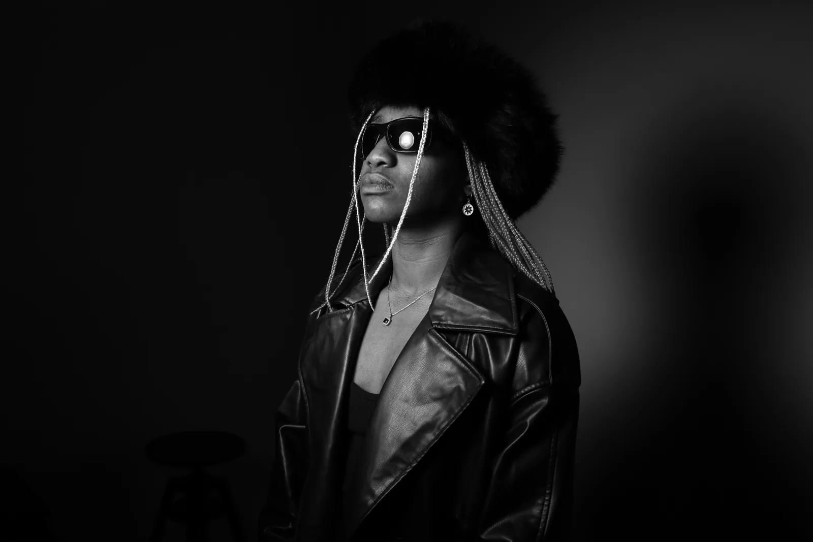 Cha La Mélo de face, buste, lunettes noires traversées d’un reflet blanc. Veste de cuir ouverte, fond gris dégradé.