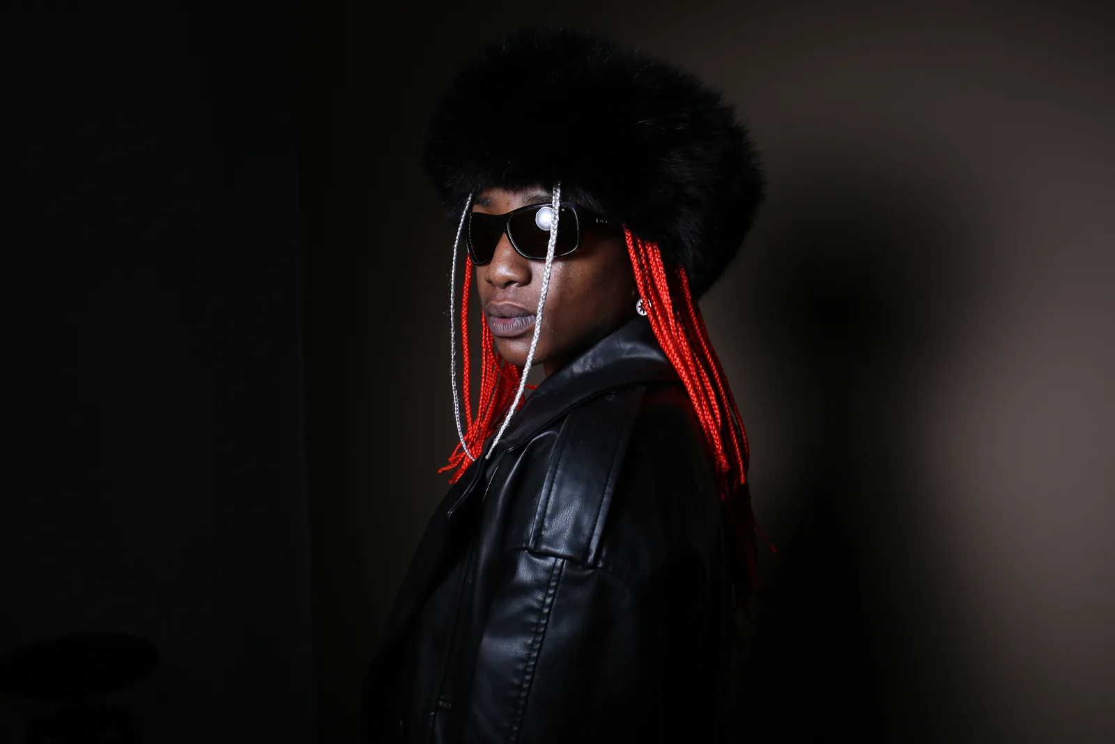 Cha La Mélo de trois quarts, lunettes noires, chapeau de fourrure et tresses rouges. Veste de cuir, fond sombre. Plan taille.