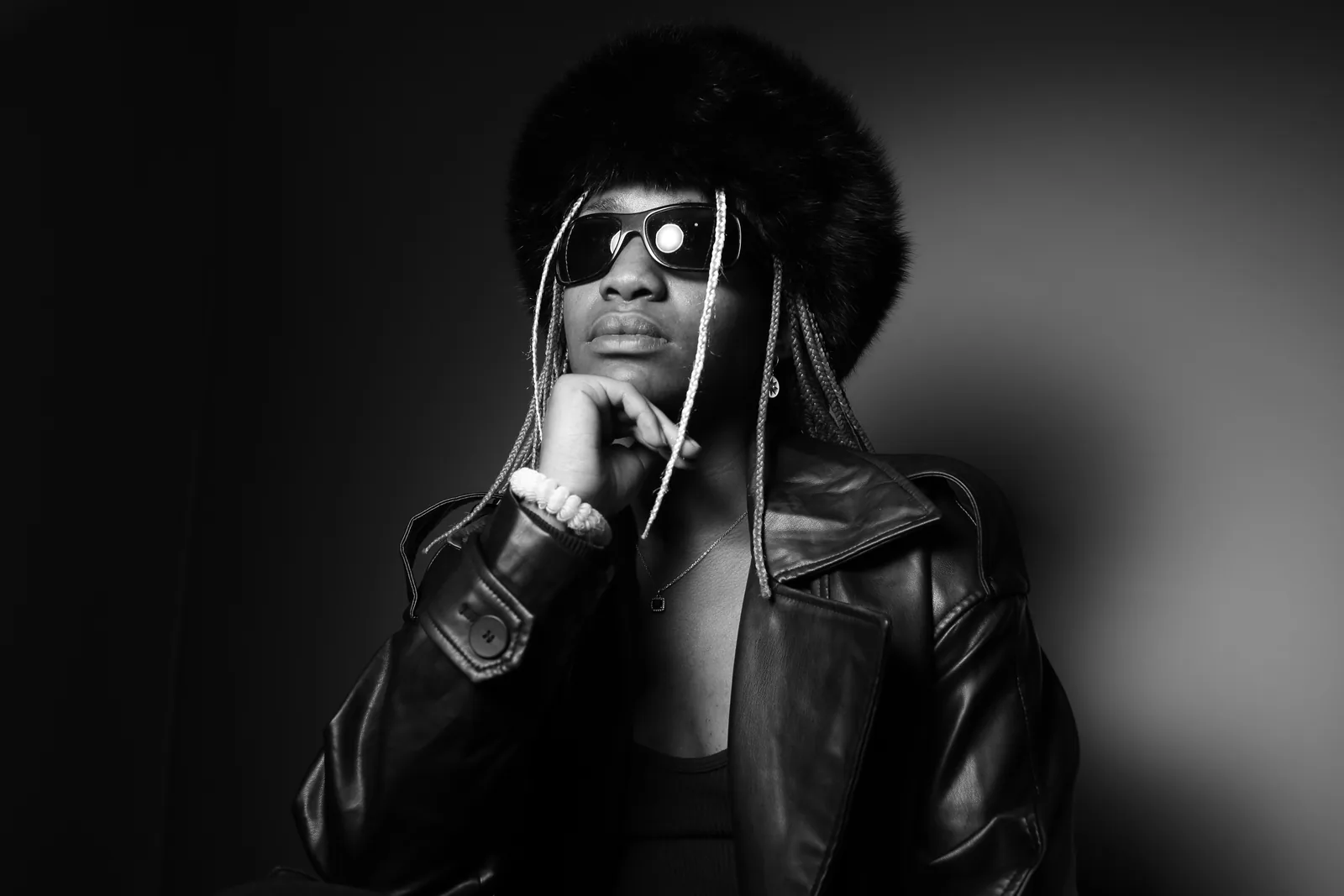 Cha La Mélo, lunettes noires et chapeau de fourrure, la main sous le menton, le regard tourné vers le haut.